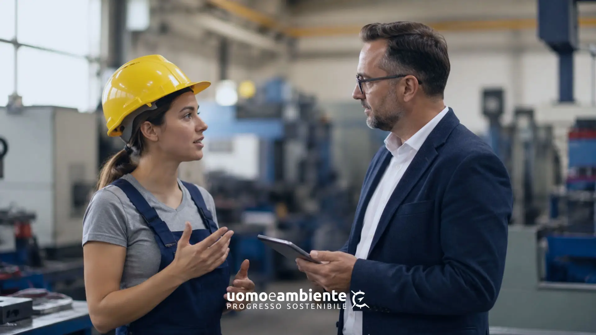 Lavoratrice in fabbrica con casco di sicurezza mentre condivide indicazioni operative con un responsabile che la ascolta.